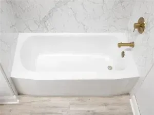 A white bathtub with gold-toned fixtures against marble-patterned walls and light wood-look flooring.