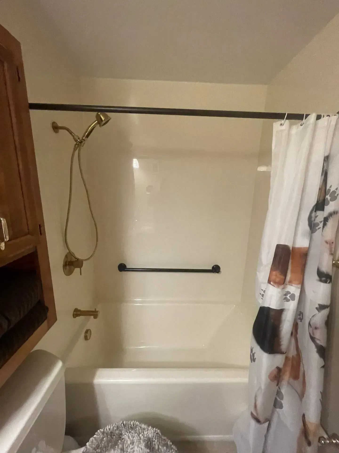 A bathroom shower and tub area with cream walls, a gold shower fixture, a black grab bar, and a patterned shower curtain.