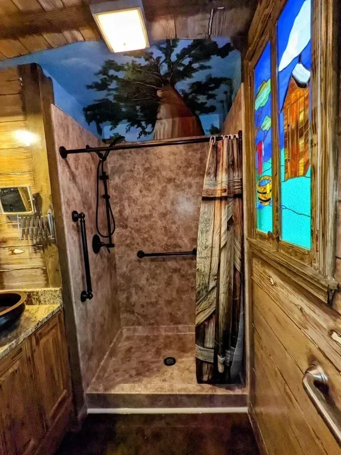 Rustic bathroom with a tile shower, grab bars, a themed shower curtain, and a ceiling mural of a tree on a blue sky.