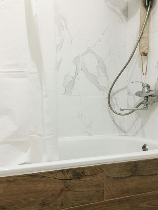 A white bathtub with marble-look wall tiles and a wooden-textured front panel, featuring a shower head and mixer tap.