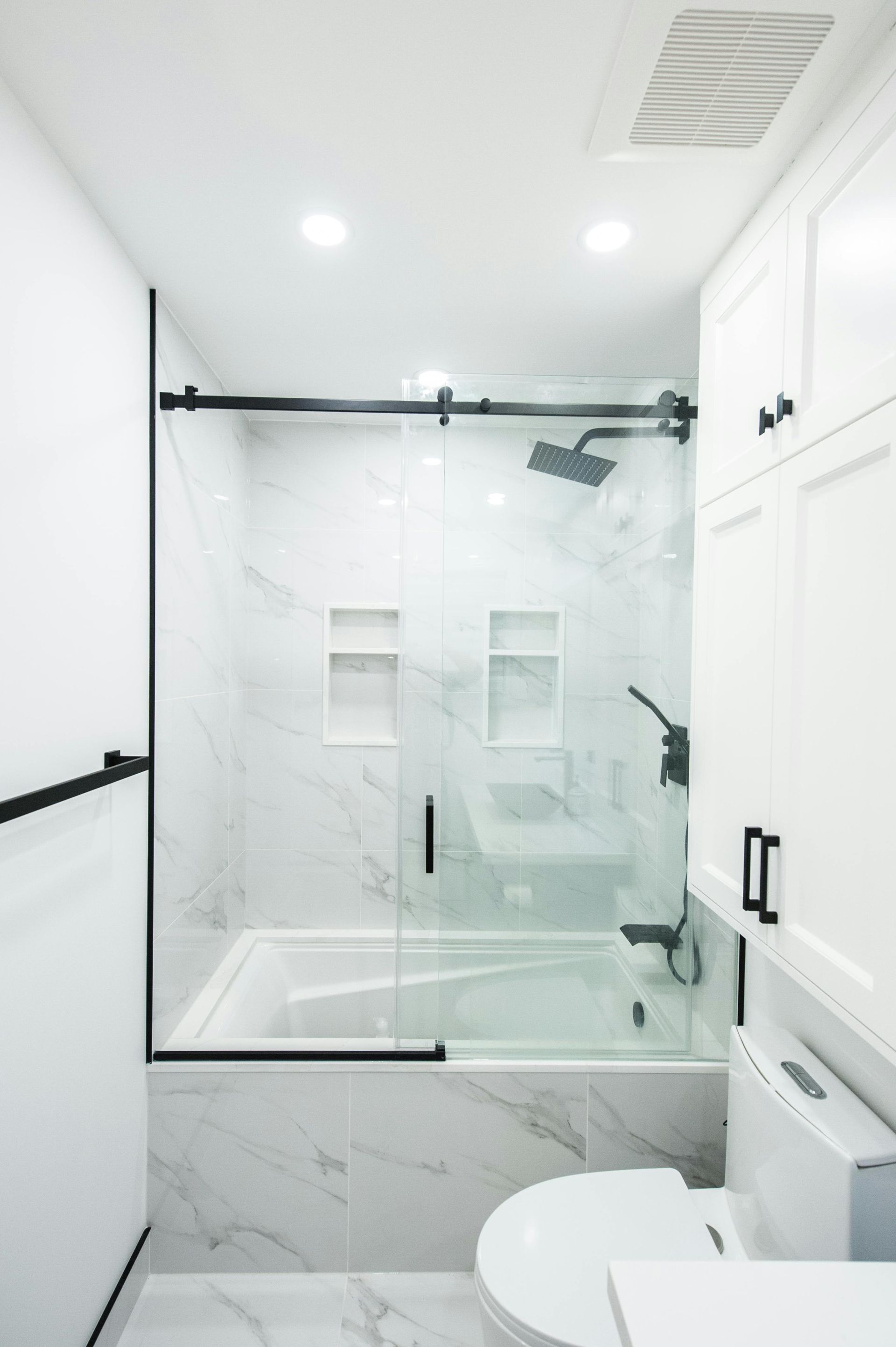 Modern bathroom with a white marble tub, glass shower enclosure, black hardware, and white storage cabinets.
