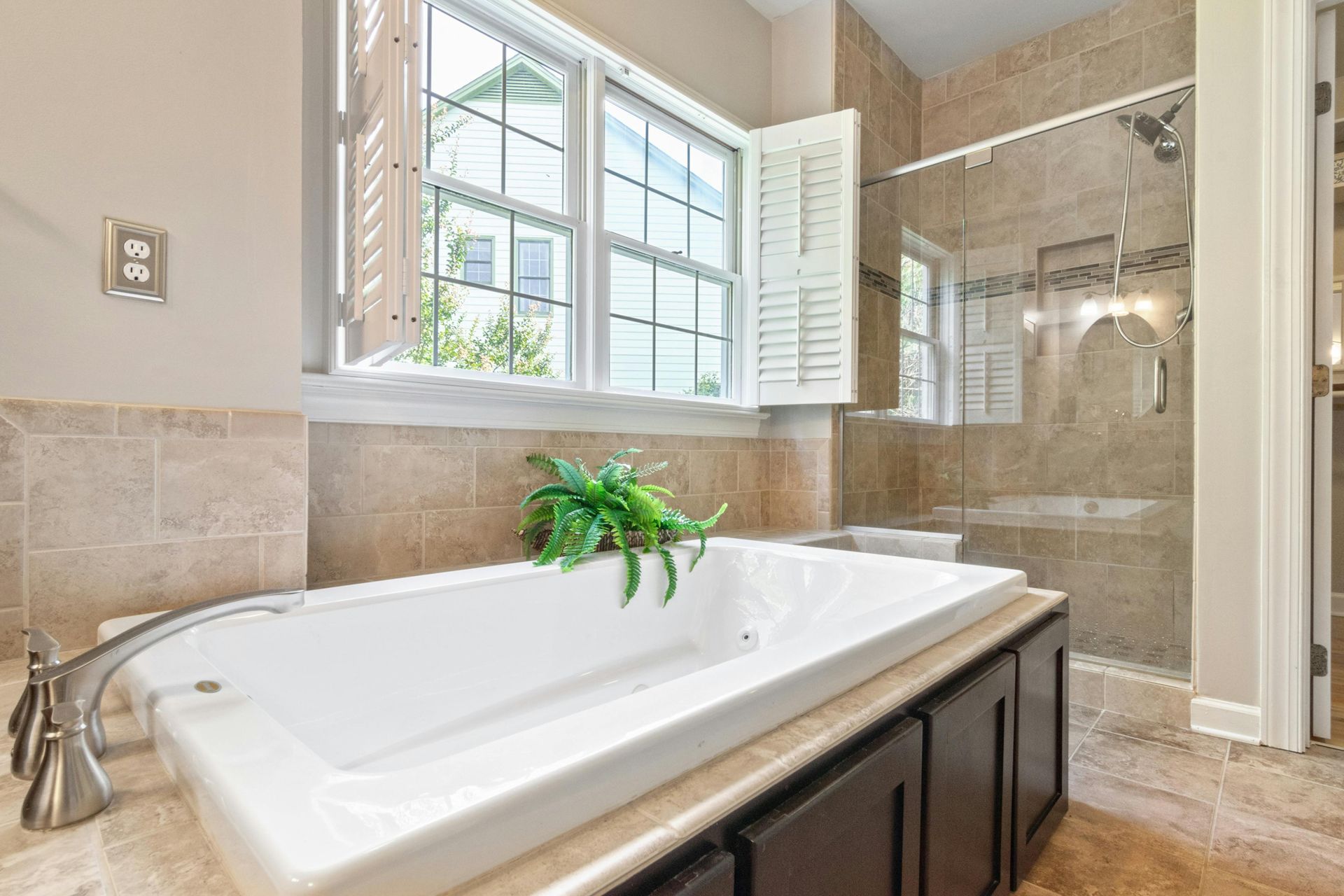 A white drop-in bathtub with a faucet and potted plant next to a window in a bathroom with a glass-enclosed shower.