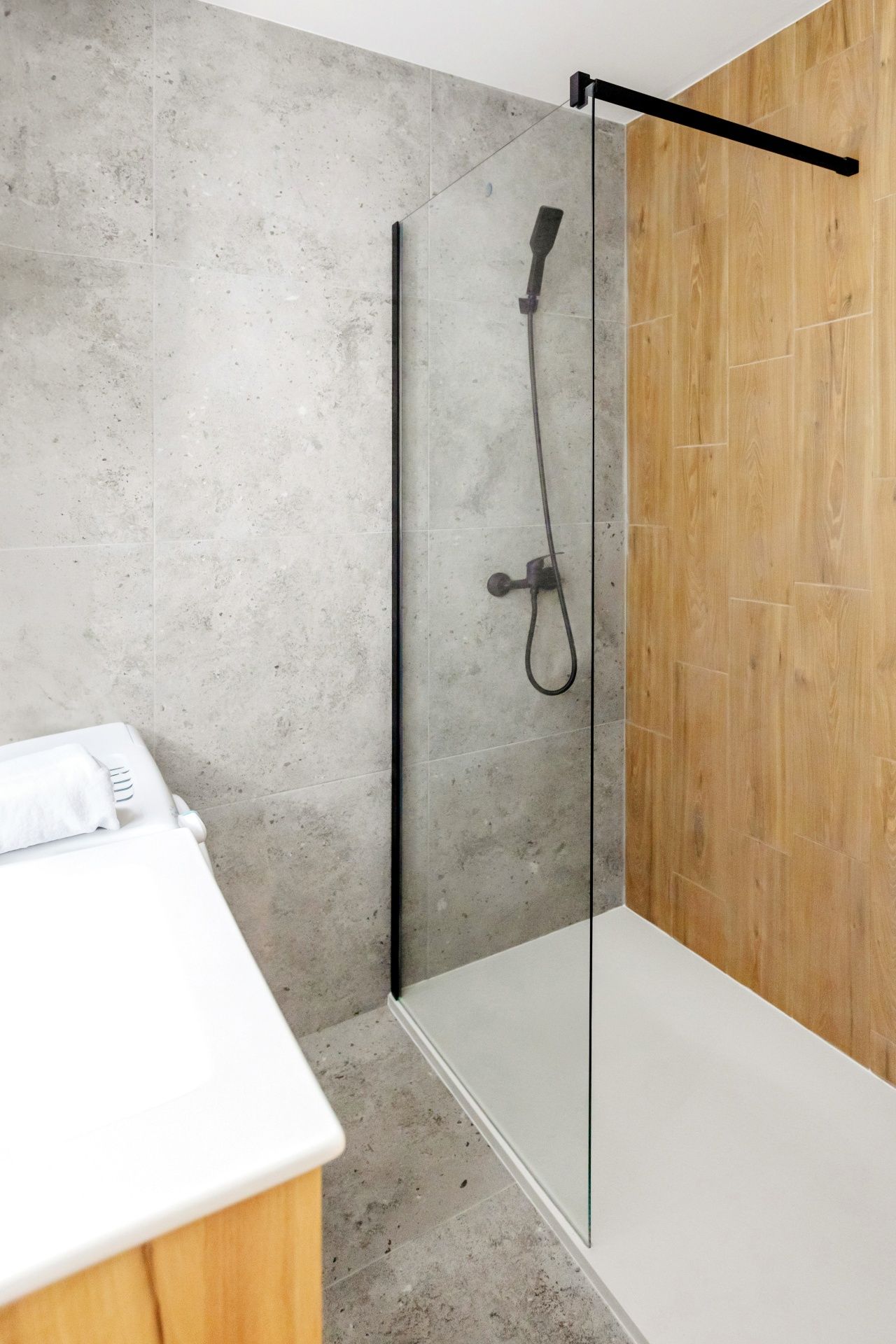 A modern bathroom with a glass-walled shower featuring a black fixture against grey concrete and wood-paneled walls.
