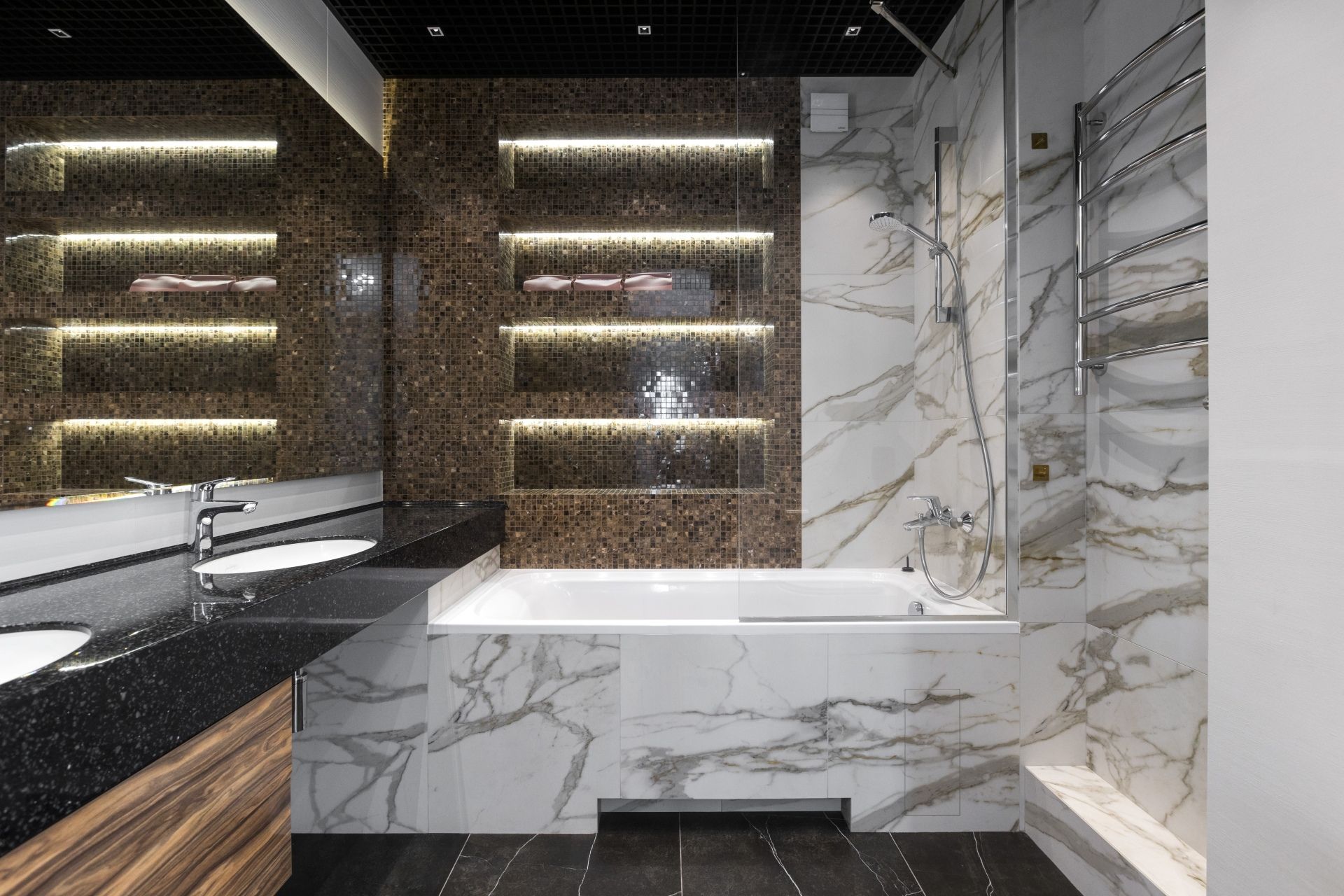 Modern bathroom with white marble-patterned bathtub and shower, brown textured shelves with lights, and a black vanity.