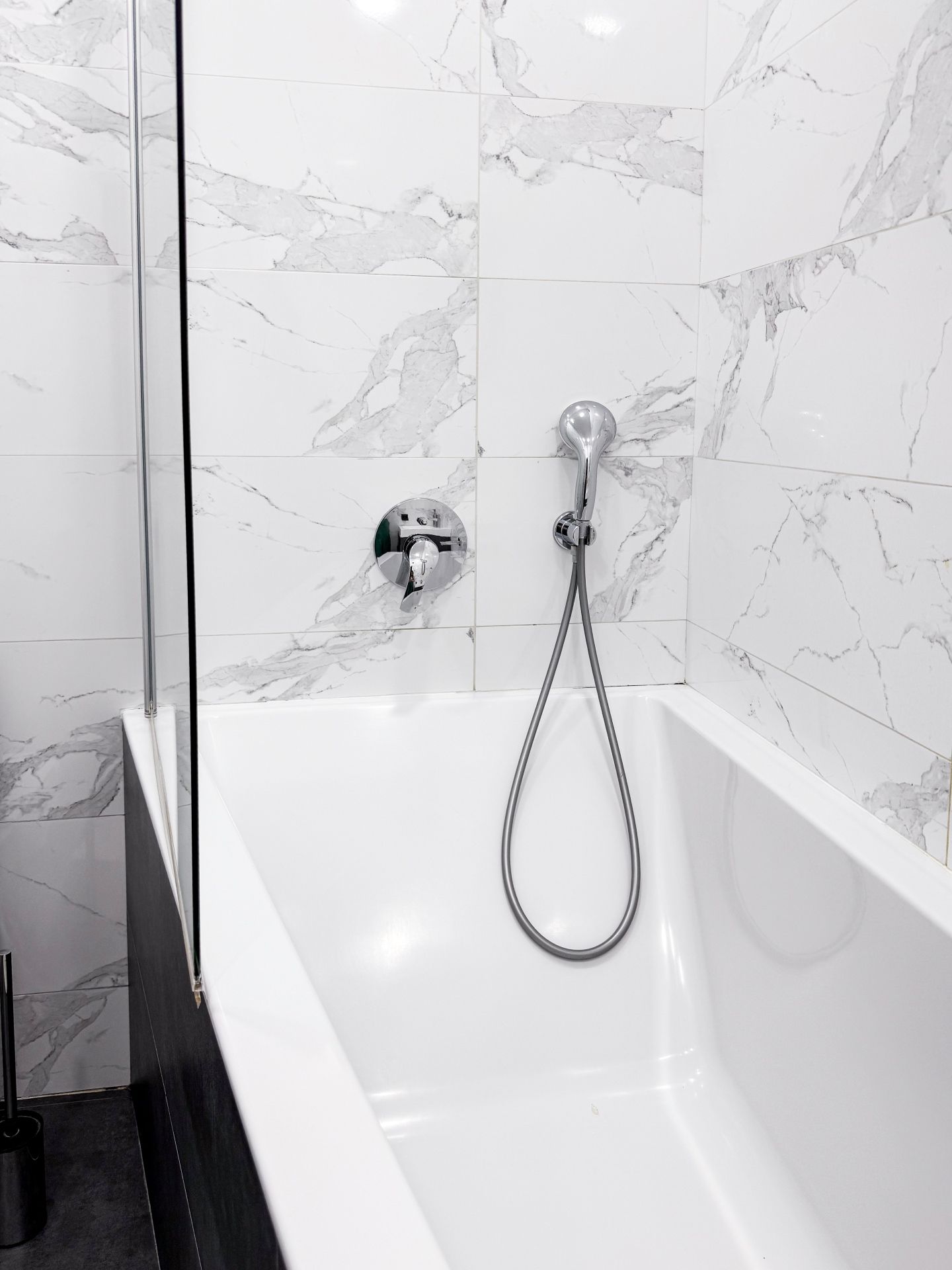 White bathtub with a handheld shower head mounted on a wall featuring white marble-patterned tiles.