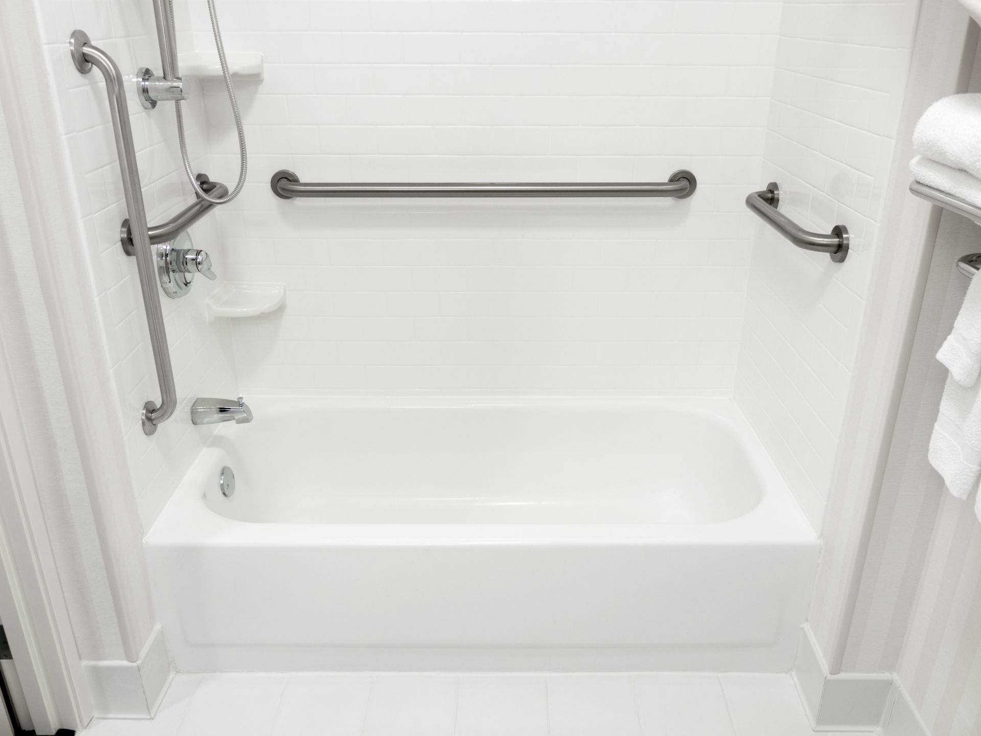A white bathtub in an accessible bathroom with multiple metal grab bars and a handheld showerhead mounted on a vertical bar.