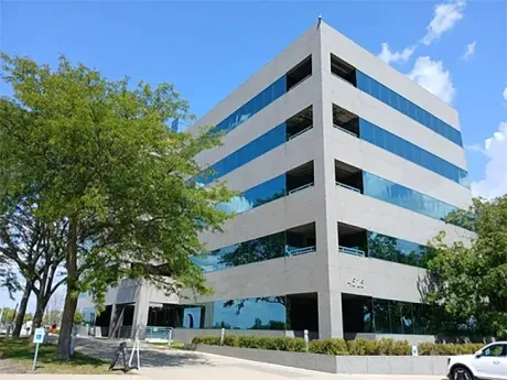 Modern white office building with blue-tinted windows beside a tree-lined street under a blue sky