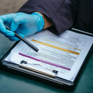 Gloved hand pointing to a highlighted medical form on a clipboard with a pen nearby