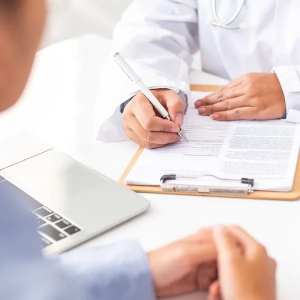Doctor writing on clipboard during a consultation beside a laptop in a bright office