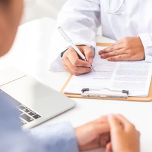 Doctor writing on clipboard during a consultation beside a laptop in a bright office