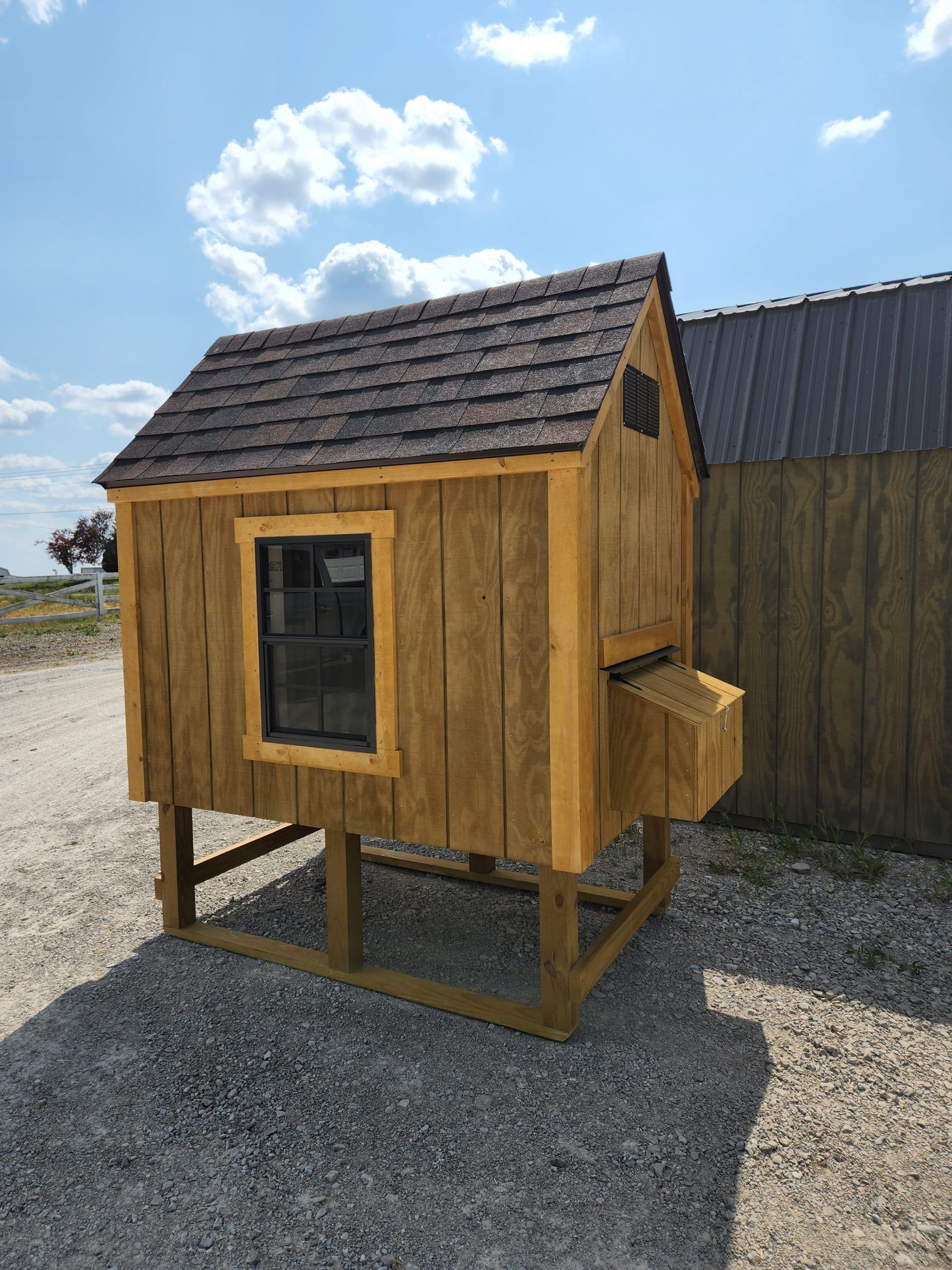 A small, wooden chicken coop with a brown shingled roof, window, and nesting box stands on a gravel surface under a blue sky.