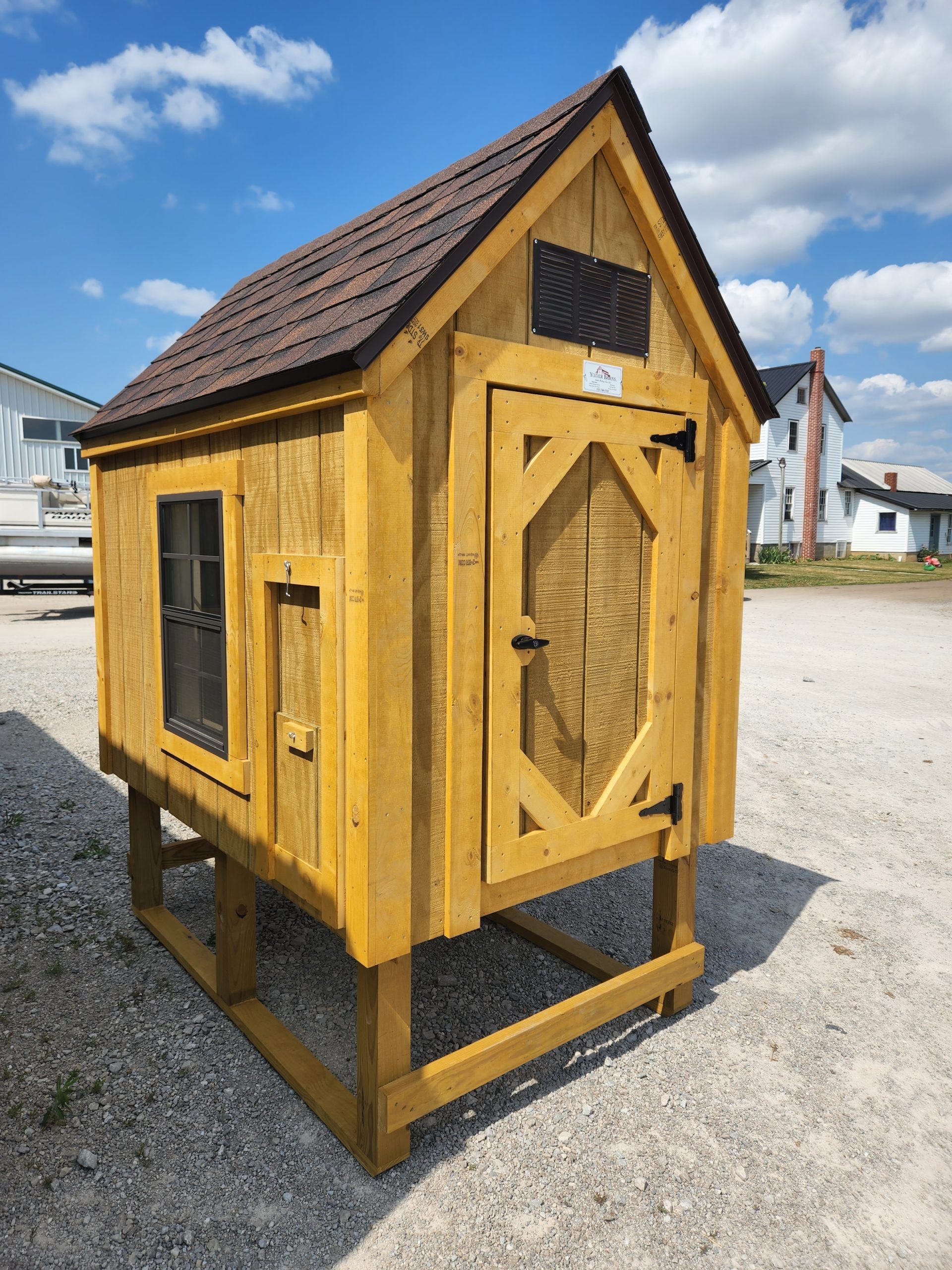 Yellow wooden chicken coop with a brown roof, sitting on a gravel lot with buildings and a blue sky in the background.