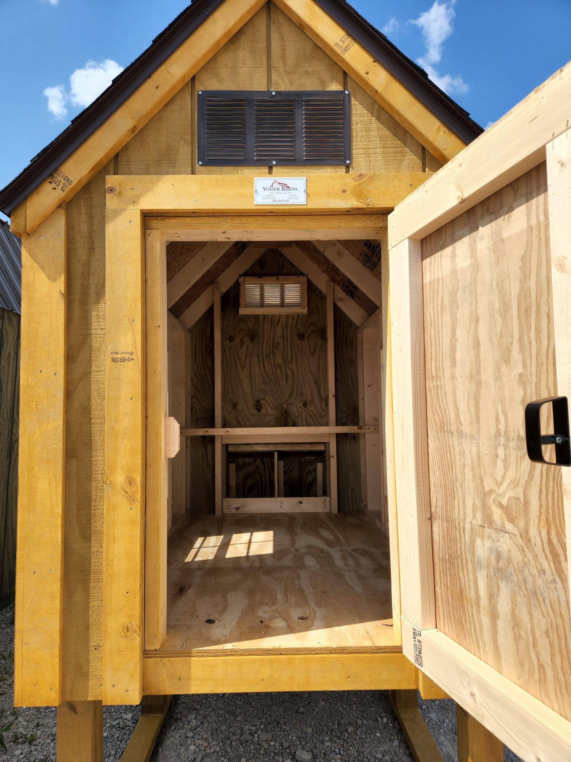 Wooden chicken coop with an open door revealing interior nesting boxes and a vent. The coop is outdoors on a sunny day.