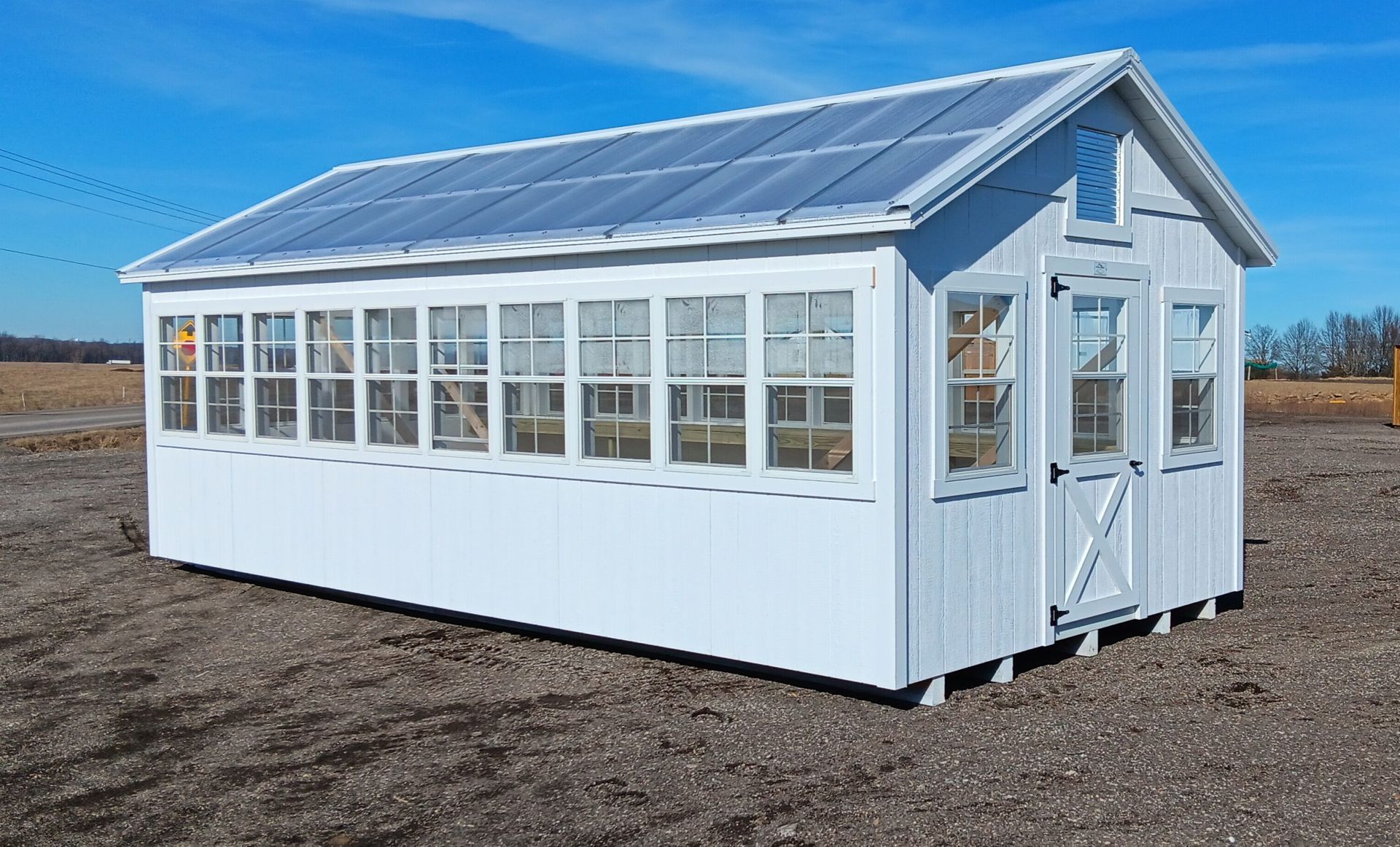 A white greenhouse with a clear roof and many windows stands on a brown field under a blue sky.