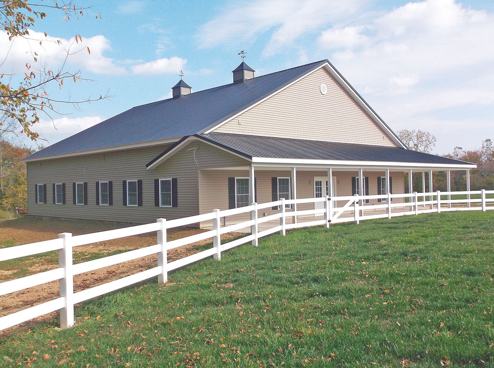 A large tan barn with a black roof and white porch, surrounded by a white fence and green grass under a blue sky.