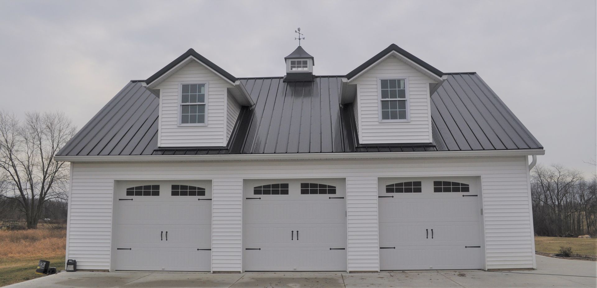 White three-car garage with black metal roof, two dormers with windows, and a small decorative cupola on a cloudy day.