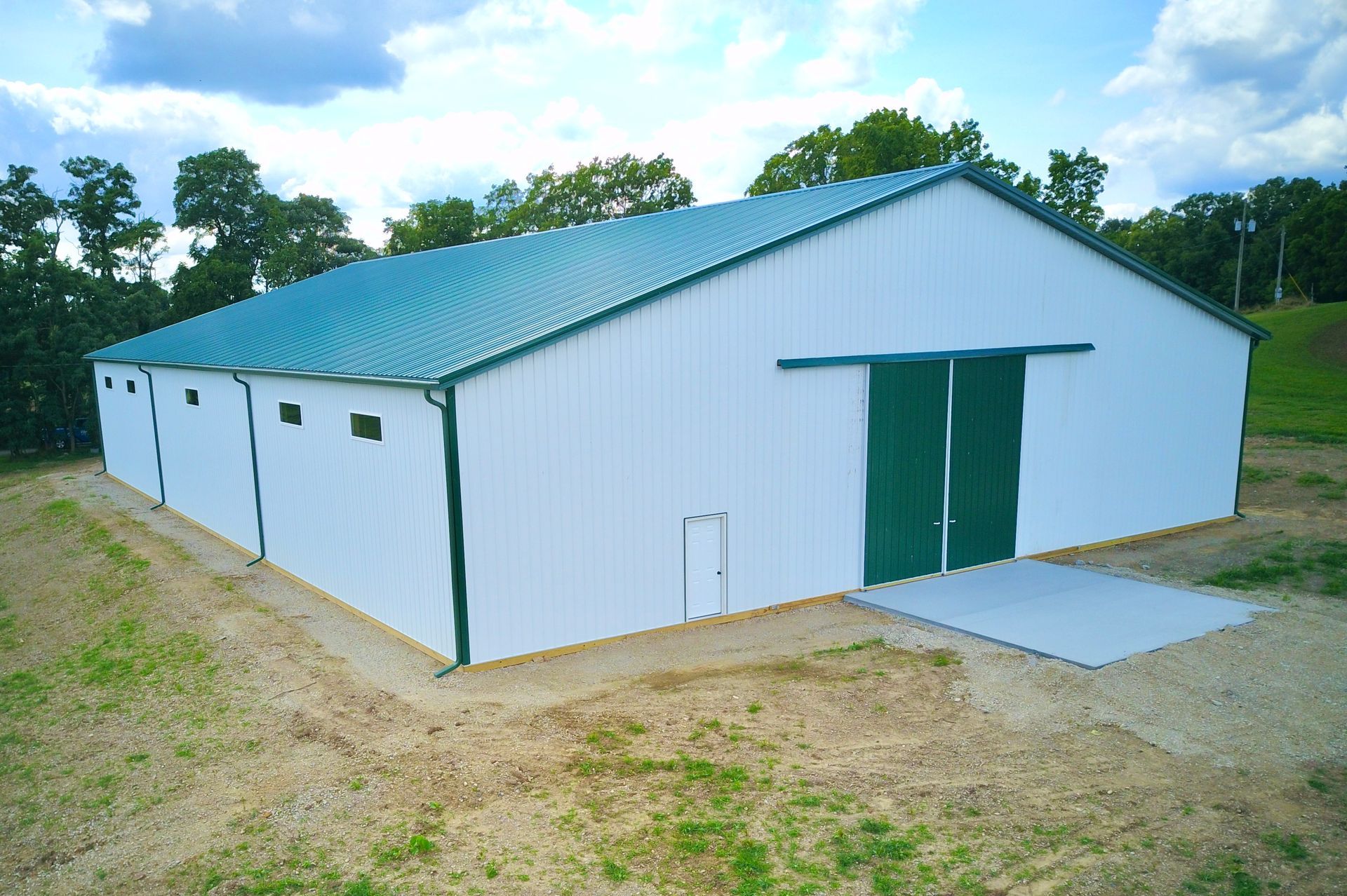 White and green metal barn with a green roof and sliding doors, set on a gravel base in a grassy area under a cloudy sky.