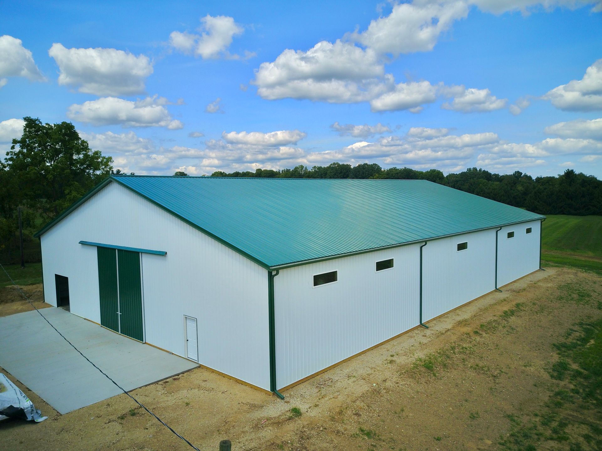 White and green metal barn under a blue sky with fluffy clouds, situated in a rural landscape.