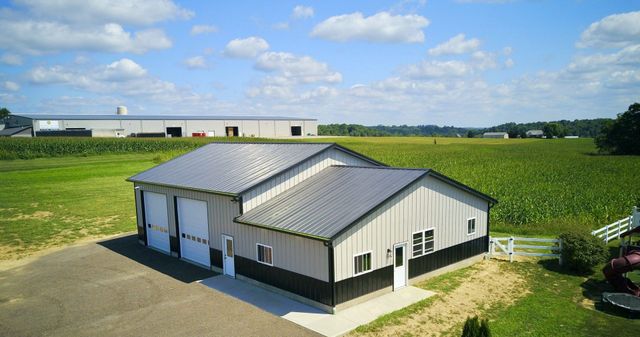 A metal building with a gray roof and white doors sits in a field, with another building visible in the background.