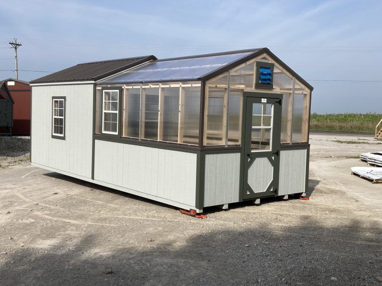 A small, pale green greenhouse with a clear roof and an attached sunroom on a gravel lot.