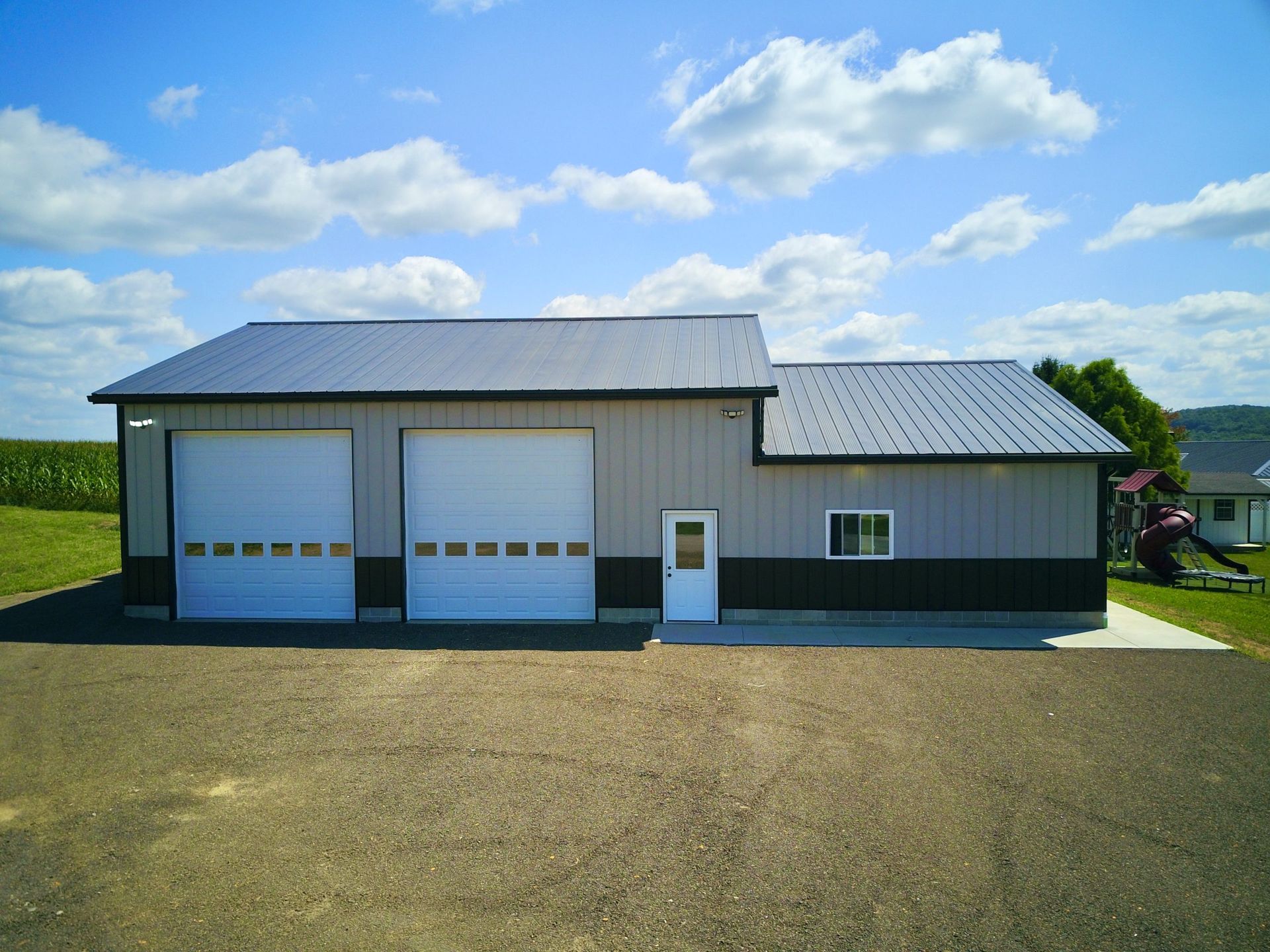 Metal-sided garage with two overhead doors, a small door, and a window under a blue sky with clouds. It's sitting on gravel.