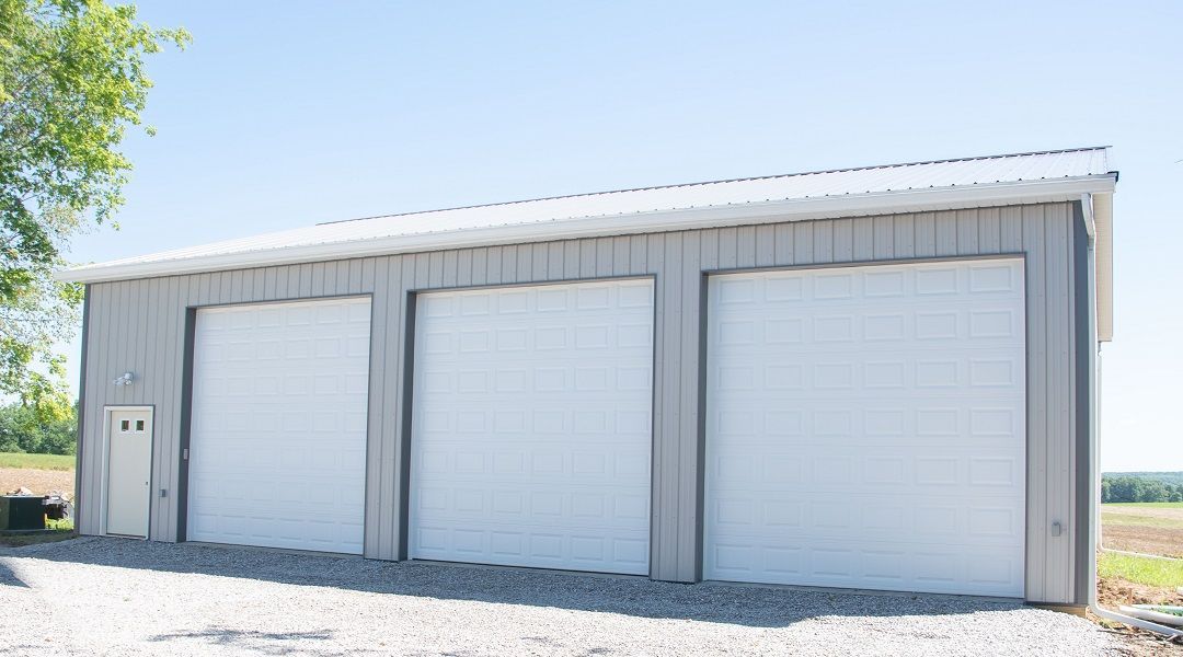 Three-bay gray metal garage with white overhead doors and a small door on the left, set against a blue sky.