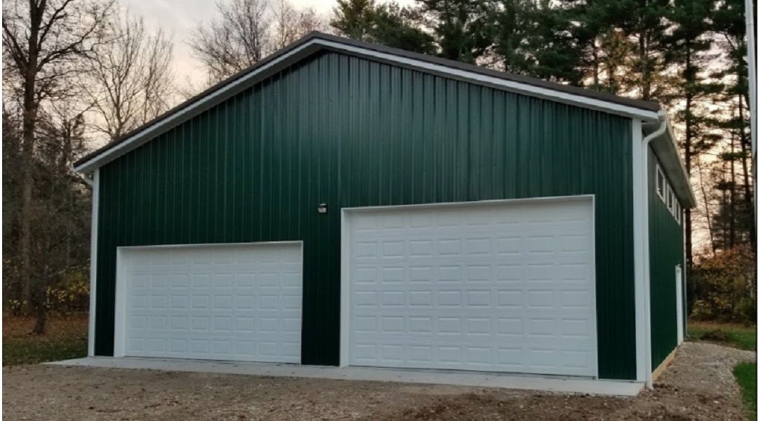Green metal garage with white garage doors and trim, on a concrete foundation, set against trees.