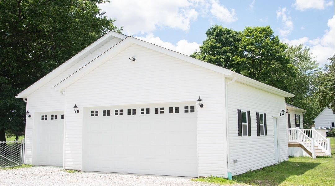 White house with a two-car garage, black accents, and a small porch. Green trees and a cloudy sky in the background.