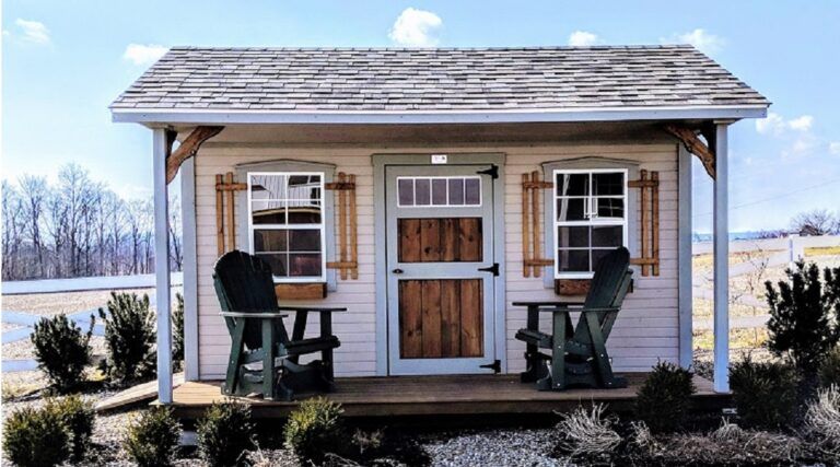 A small, tan shed with a wooden porch, featuring a brown door, two green Adirondack chairs, and wooden shutters.