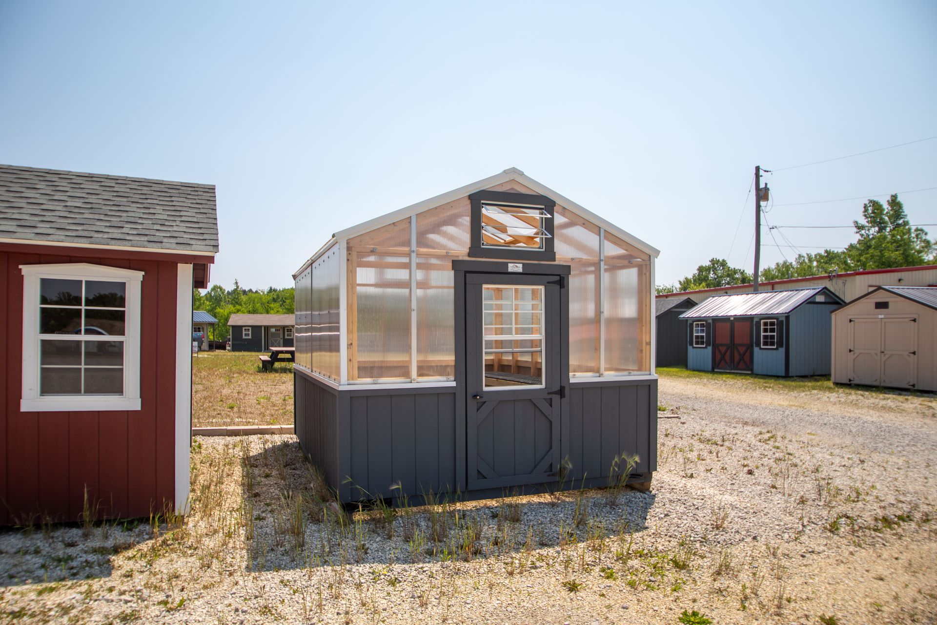 A small greenhouse with a gray base and clear plastic walls stands on gravel.