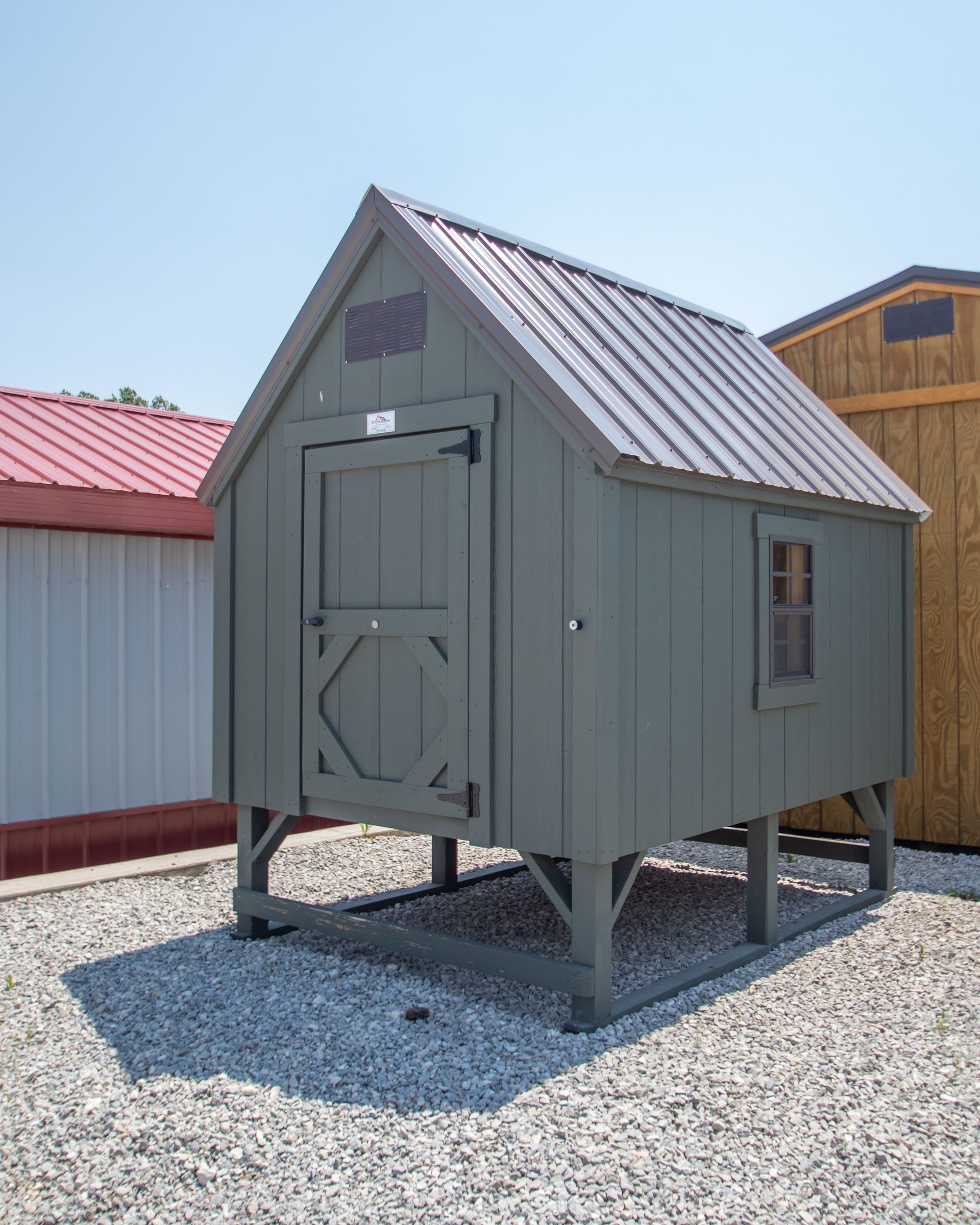 A green chicken coop with a metal roof and a small window, raised on stilts, sitting on gravel.