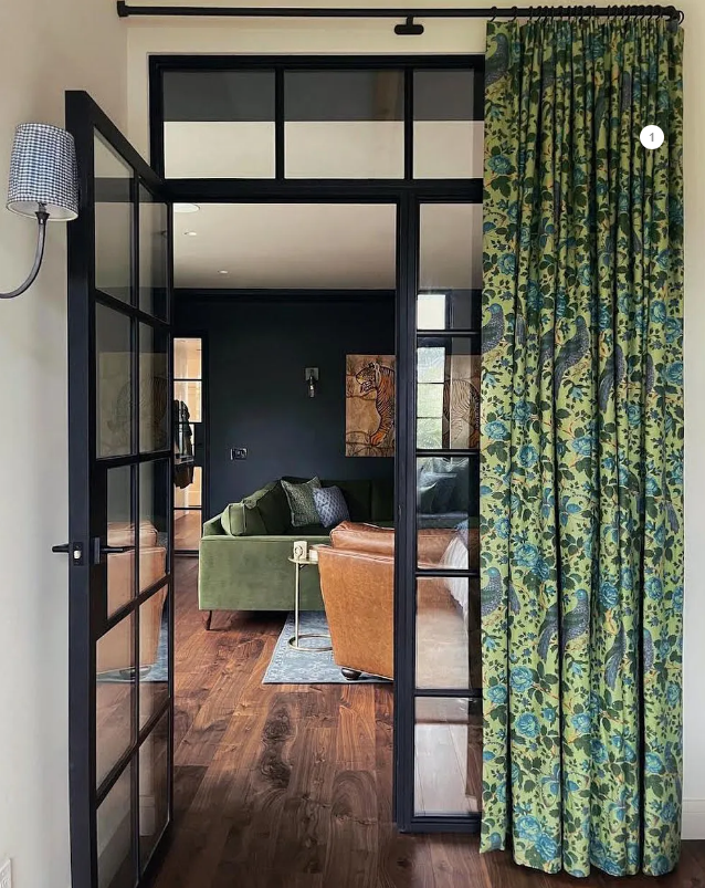 A room with dark green floral curtains, steel doors, and a view of a living room with a green sofa.