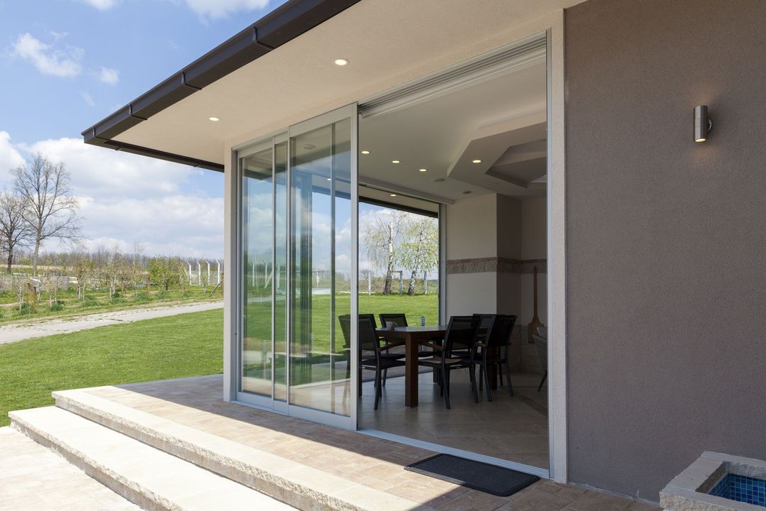 Sliding glass doors open to a dining area with a table and chairs, on a patio with steps and lawn.