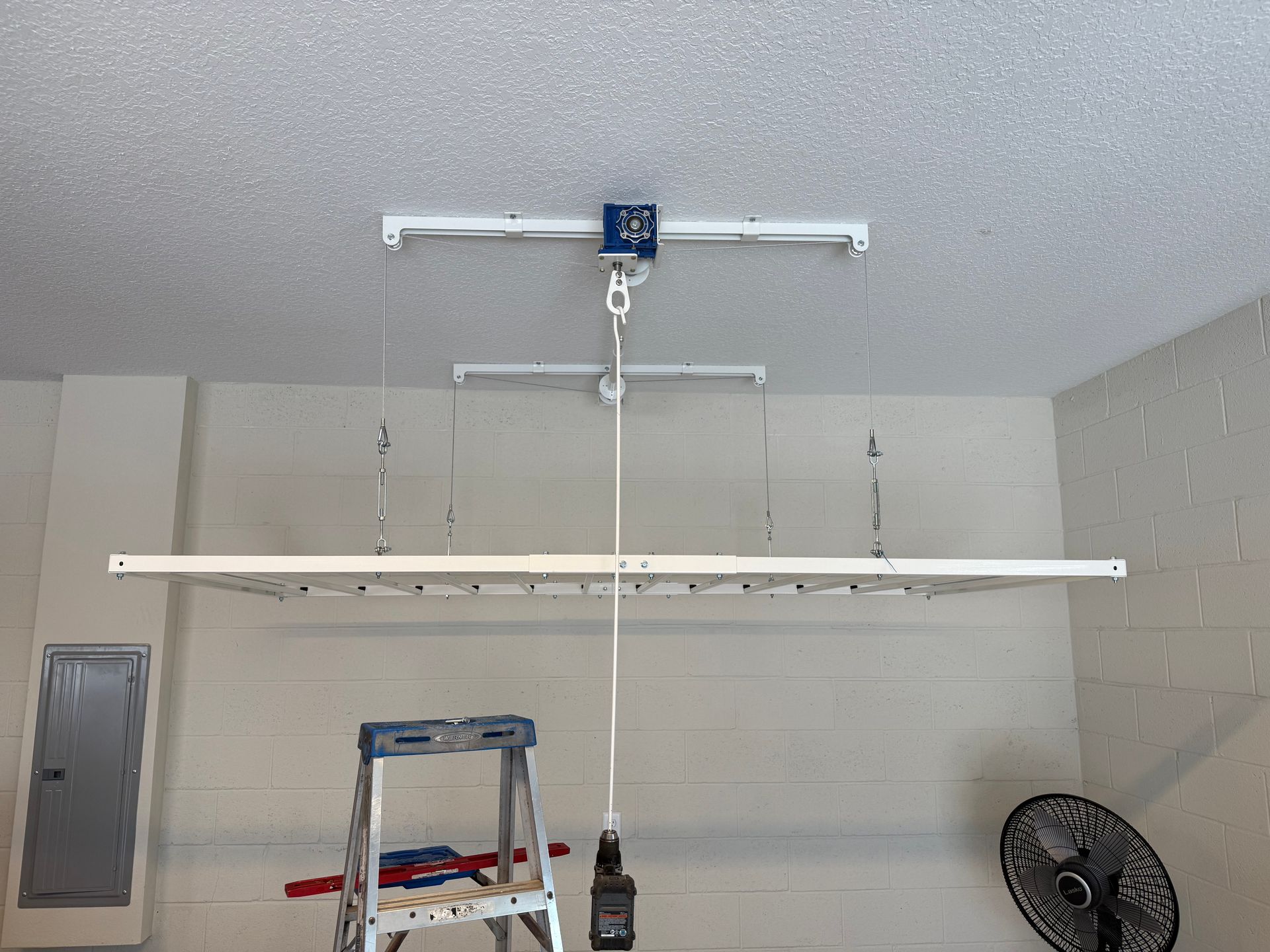 Garage ceiling with a mounted garage door opener, hanging rails, ladder, and a standing fan below.