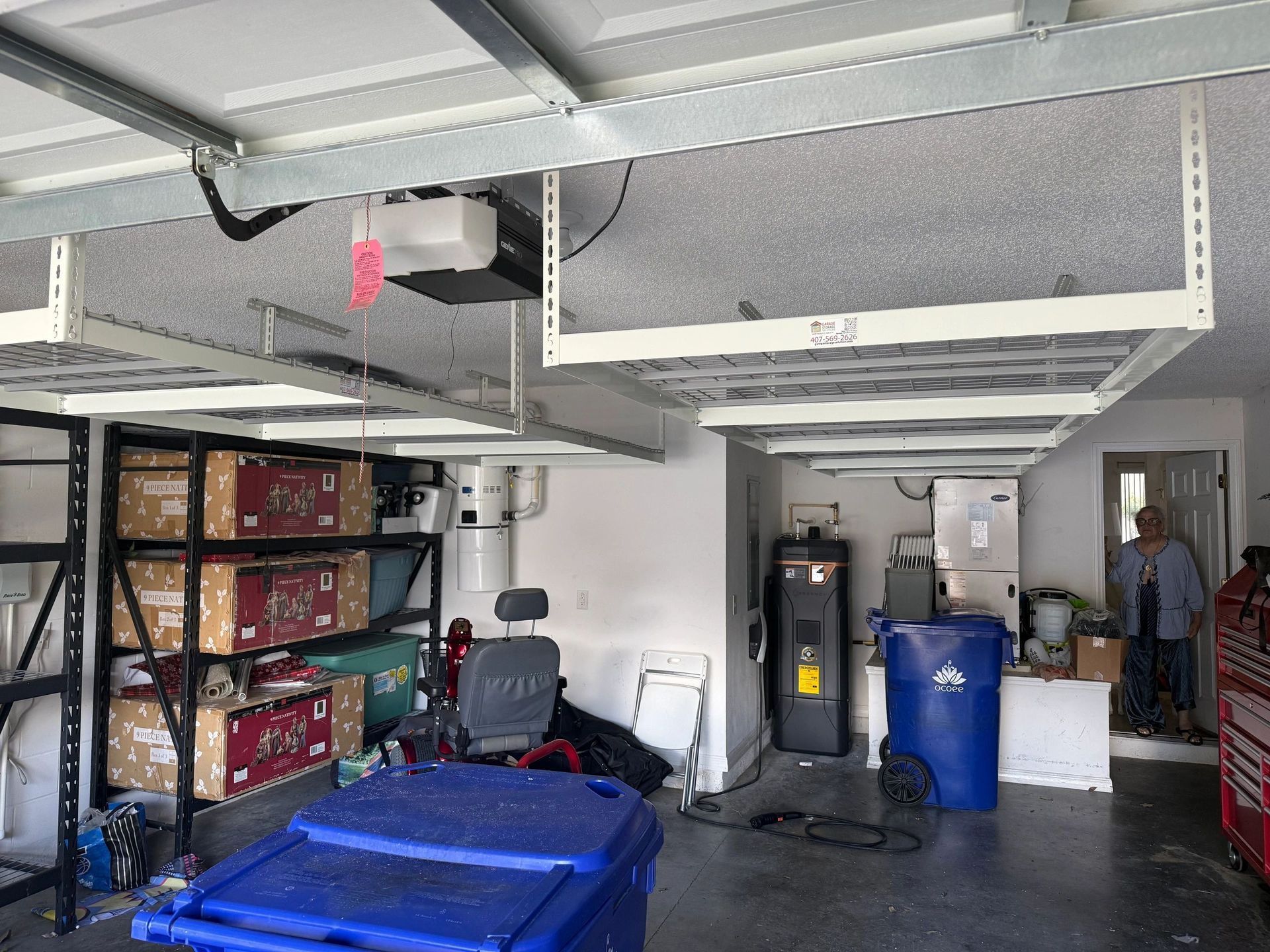 Garage interior with shelving, stacked boxes, blue barrels, and parked vehicles under a ceiling-mounted lift.