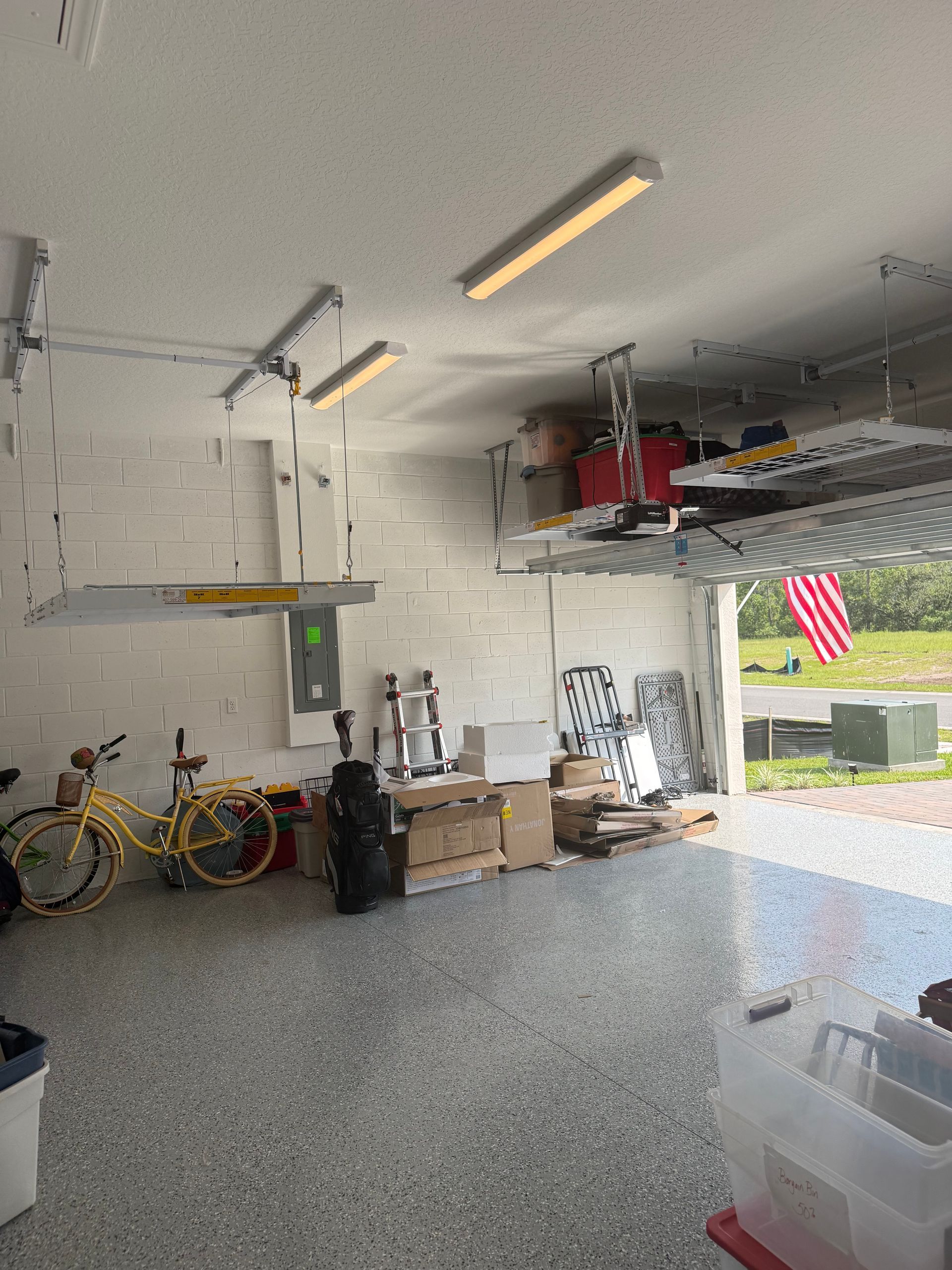 Garage with bicycles, storage items, and overhead lights; open door shows driveway and American flag outside