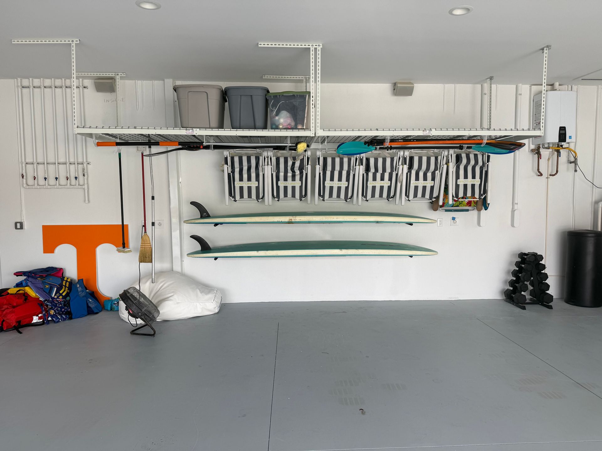 Garage storage wall with organized sports equipment, shelves, hanging tools, and stacked cushions under white cabinets