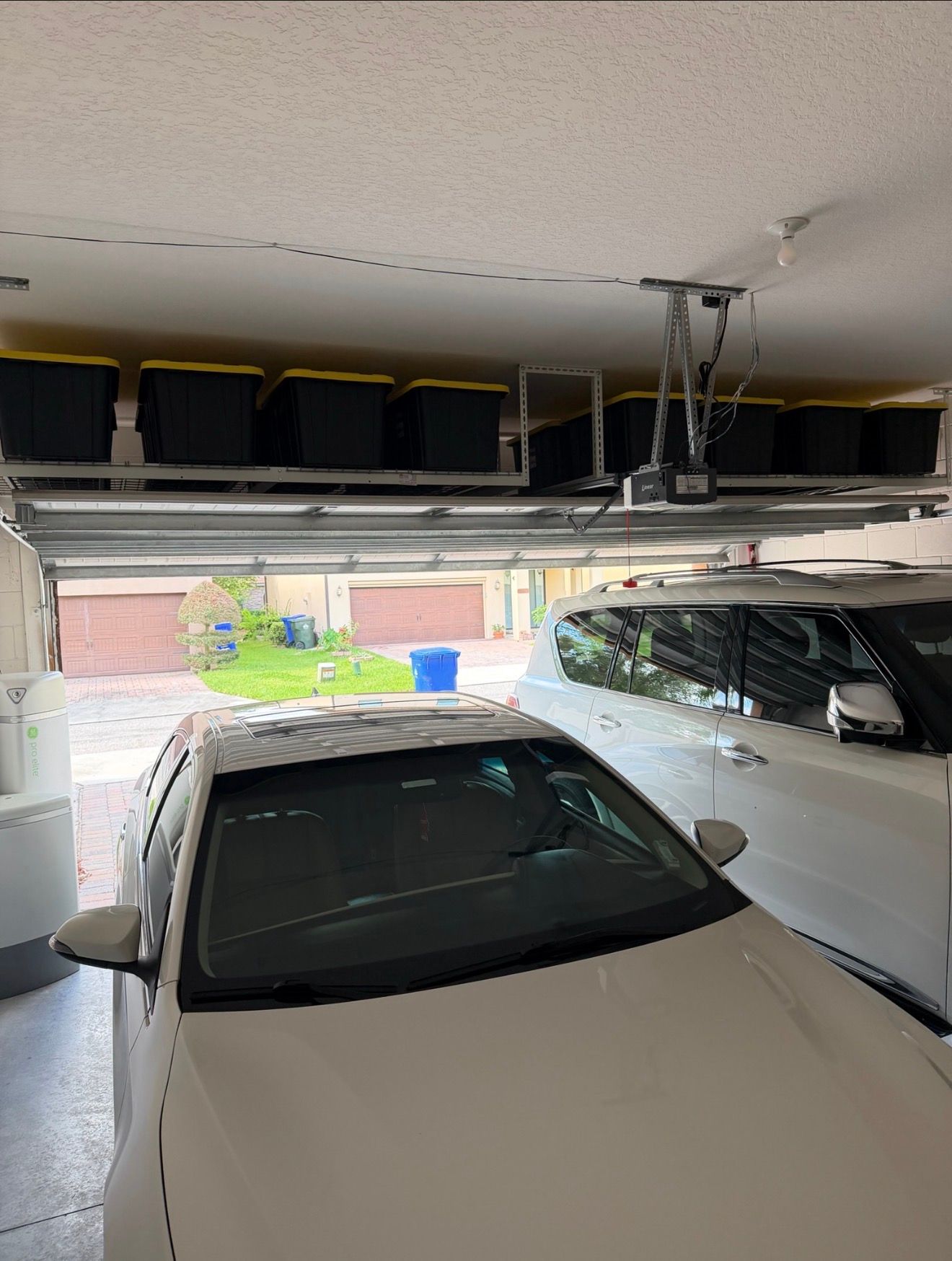 Two cars parked in a garage with the door open, showing storage bins and daylight outside.