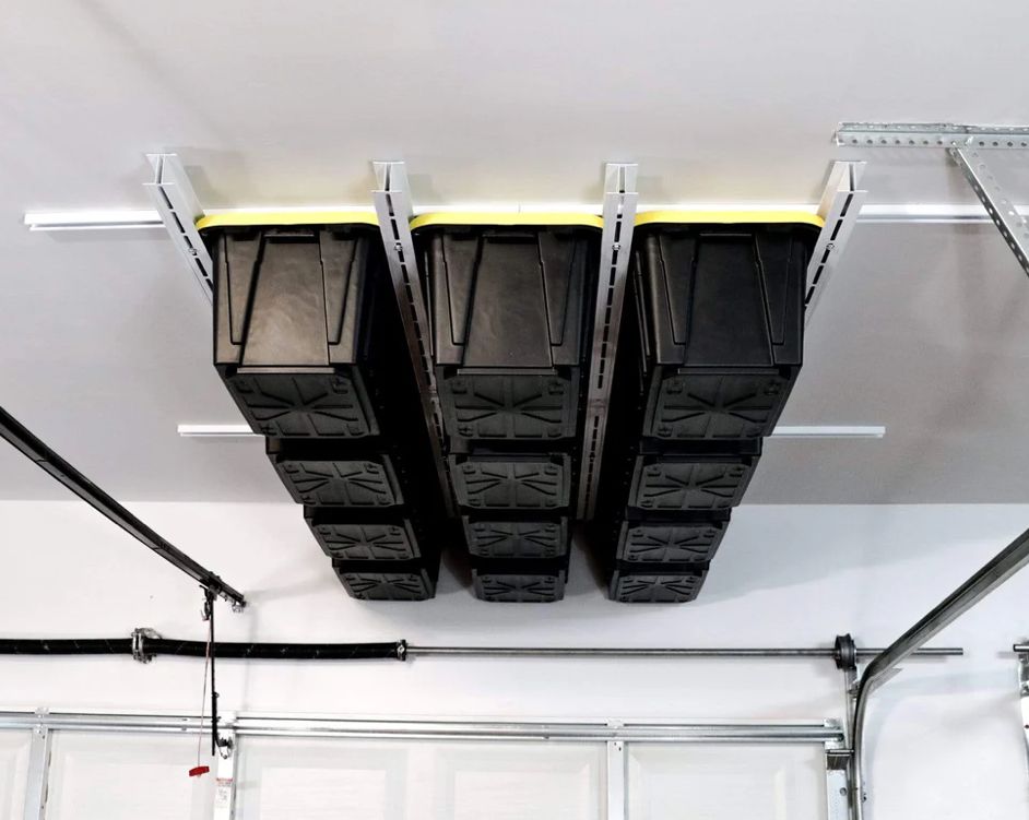 Three black storage bins mounted upside down on the garage ceiling with yellow rails and white walls below