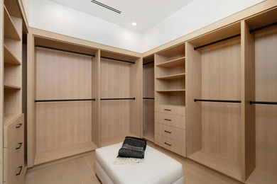 Spacious walk-in closet with light wood shelves, hanging rods, drawers, and a white ottoman.