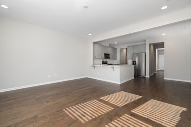 Empty modern living room with dark wood floors and white walls, open kitchen in back.