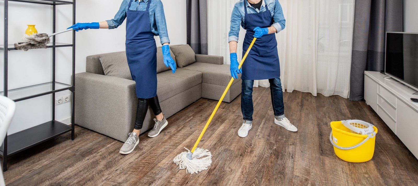 Worker in orange overalls mopping a floor beside a blue bucket near a glass wall.