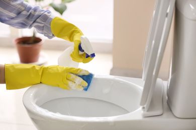 Person in yellow gloves cleaning a toilet with spray bottle and cloth in a bathroom
