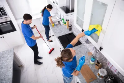Three people cleaning a bright kitchen, one wiping the backsplash and others sweeping and scrubbing the counter.