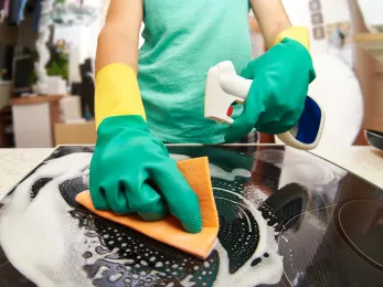 Person cleaning a black stovetop with a sponge and spray bottle, wearing green gloves and yellow sleeves