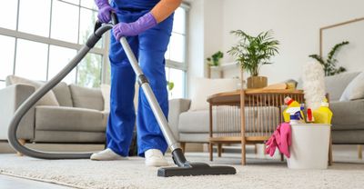 Person vacuuming a living room carpet beside a couch, chair, plants, and cleaning supplies.