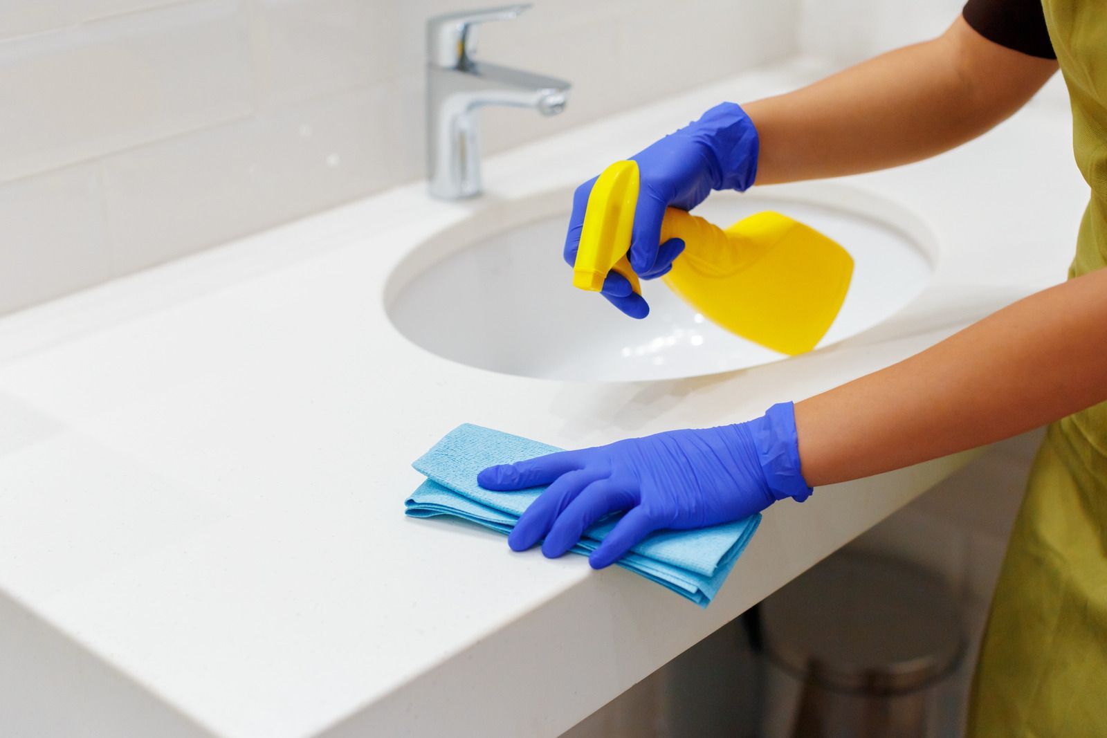 Gloved hands cleaning a white bathroom sink with a spray bottle and blue cloth