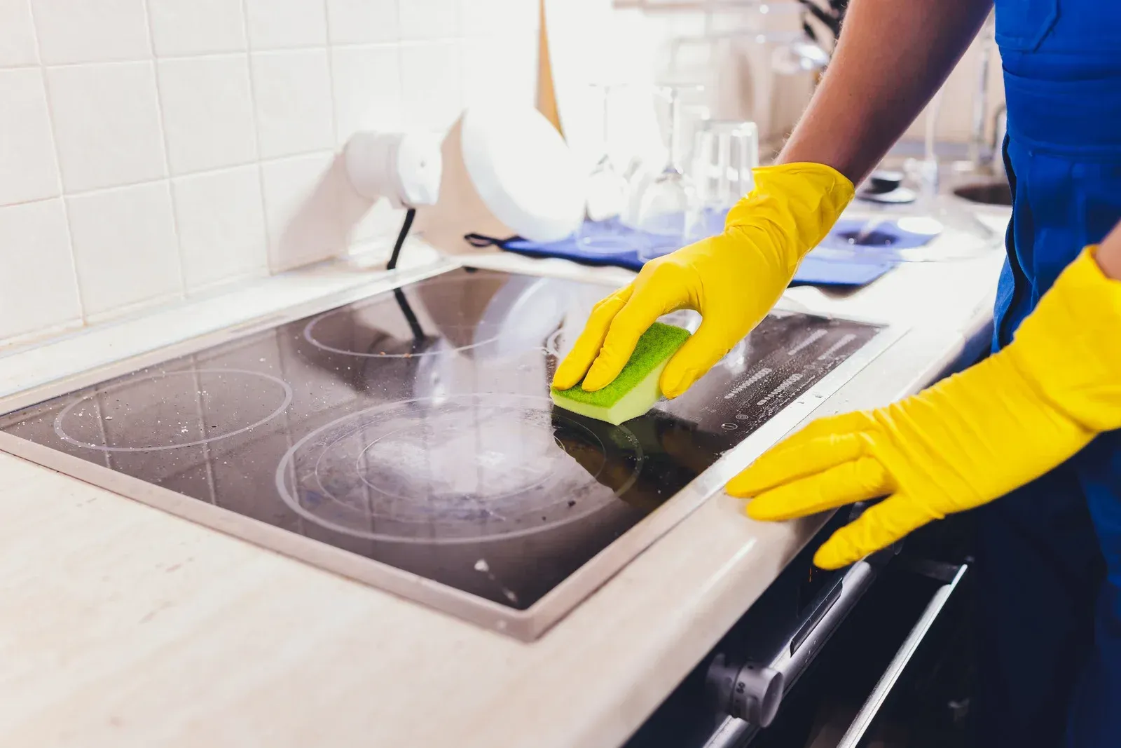 Person in yellow gloves scrubbing a glass stovetop with a sponge in a kitchen