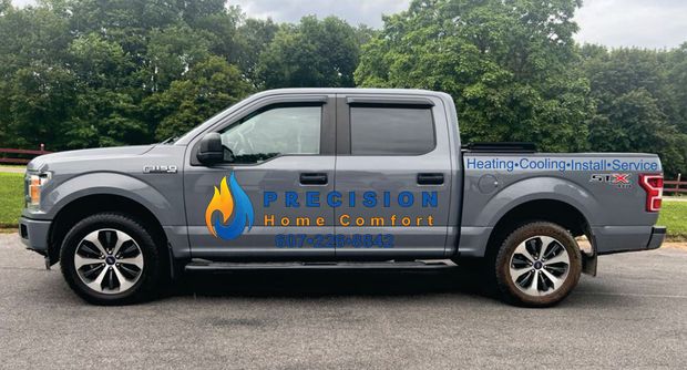 A grey Precision Home Comfort pickup truck parked outdoors in front of a tree line.