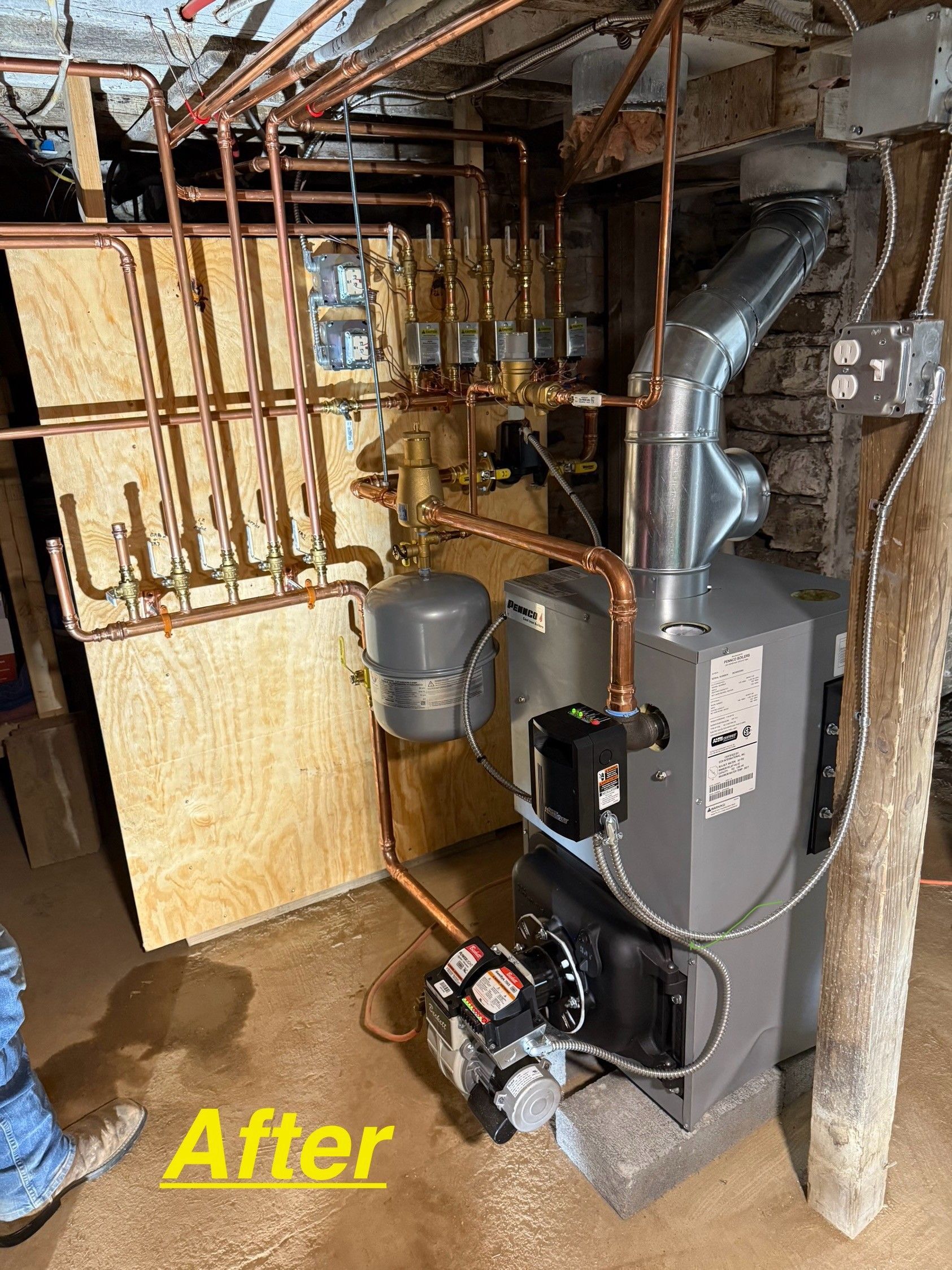 A gray residential oil-fired boiler with a new copper piping manifold and expansion tank mounted on a plywood backboard.